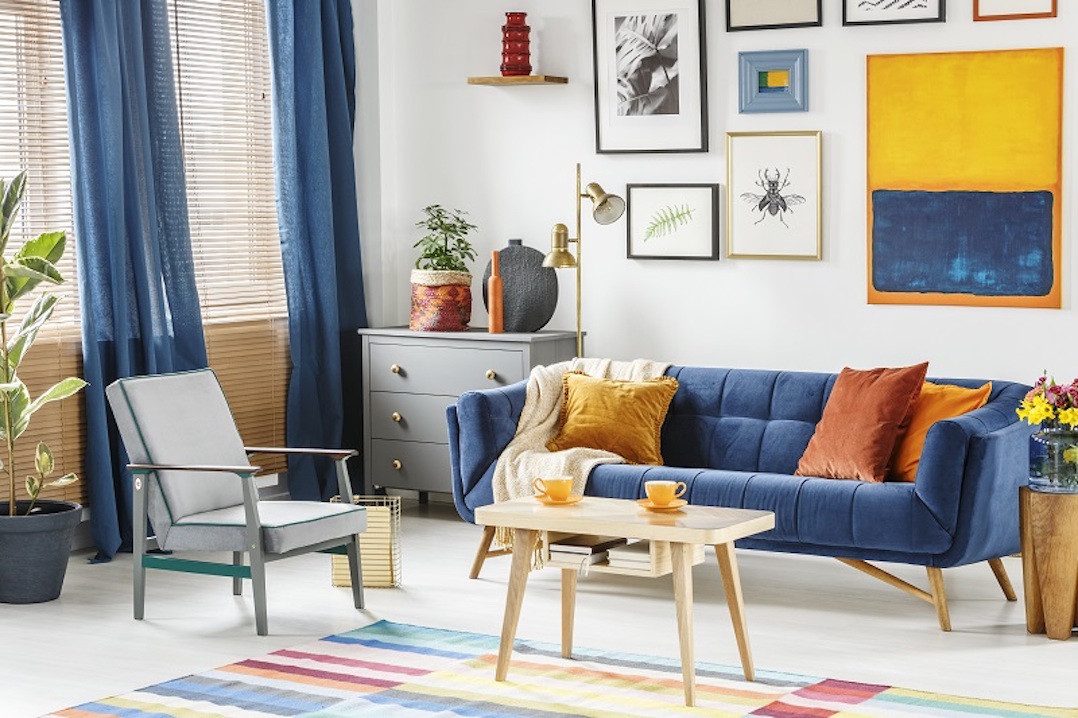 Bright and cozy living room interior with blue drapes, a sofa with orange cushions, gray armchair and a wooden table with two cups of coffee. Real photo